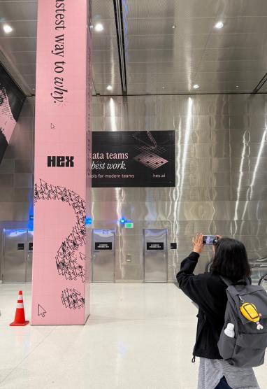 Person photographing a Hex out-of-home ad — a tall pink column with "fastest way to apply" and Hex branding — in an airport or transit hall