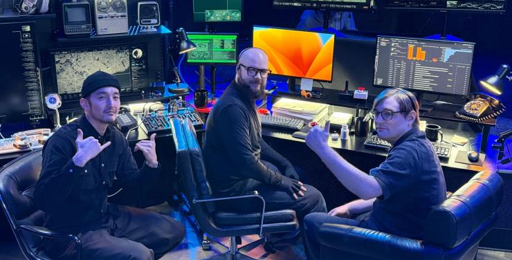 Three Hex team members posed in a dramatically lit room filled with multiple monitors, vintage tech gear, and audio equipment
