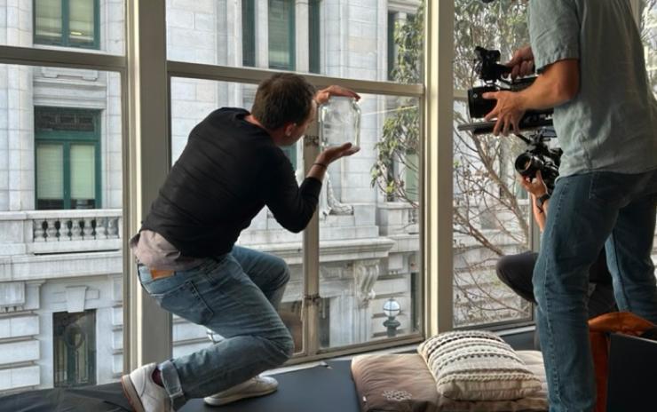 Behind-the-scenes video shoot: a person poses at a large window overlooking a city building while a camera operator films nearby