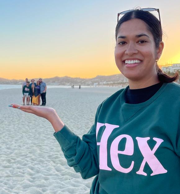 Hex team member in a green Hex sweatshirt on a beach at sunset, playfully holding tiny teammates in her outstretched palm